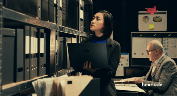 A female IT professional holding a laptop inspects shelves of physical file binders, representing the tedious and manual approach to regulatory compliance.